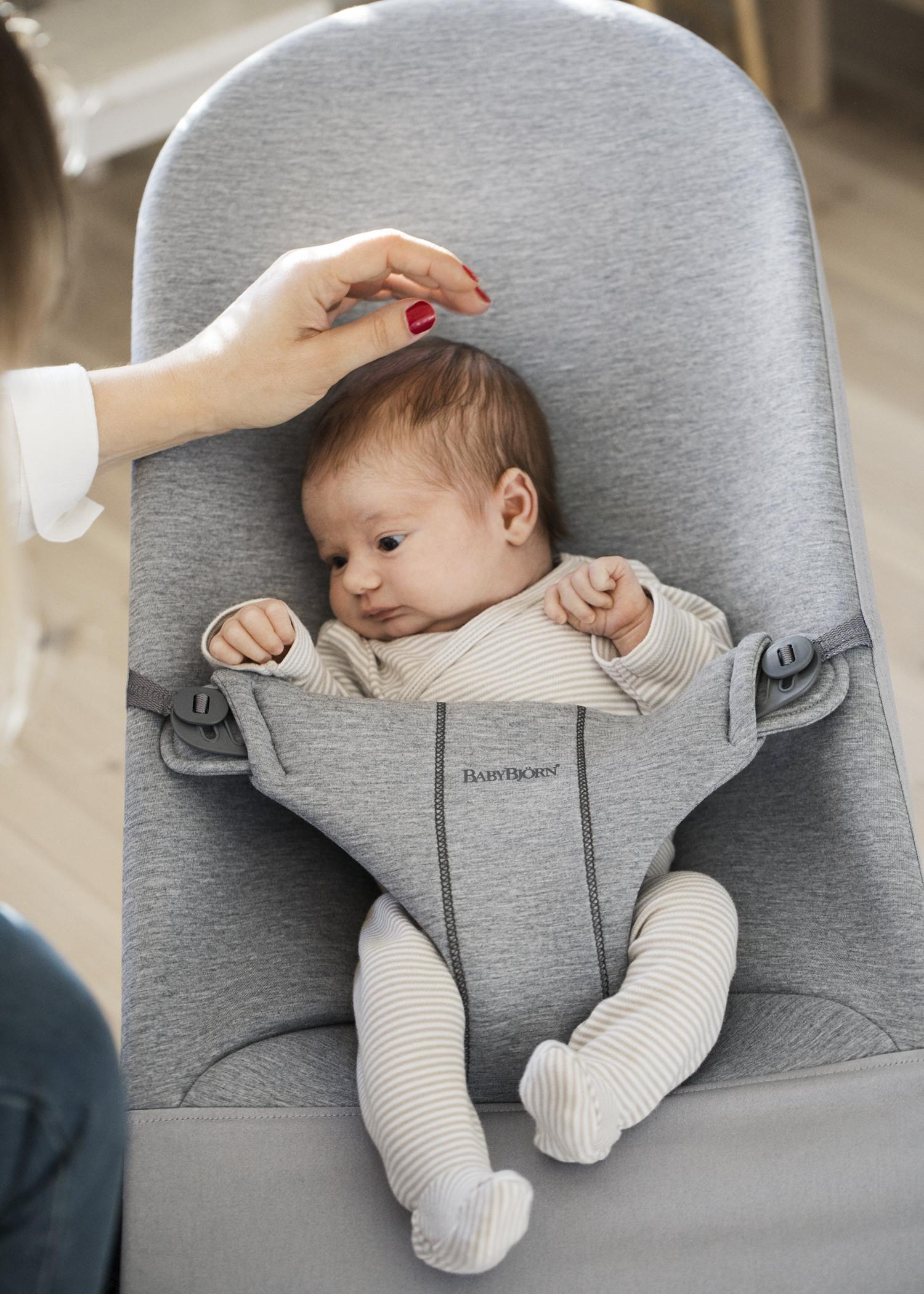 A baby in a gray bouncer wearing a striped outfit. An adult's hand gently touches the baby's head.