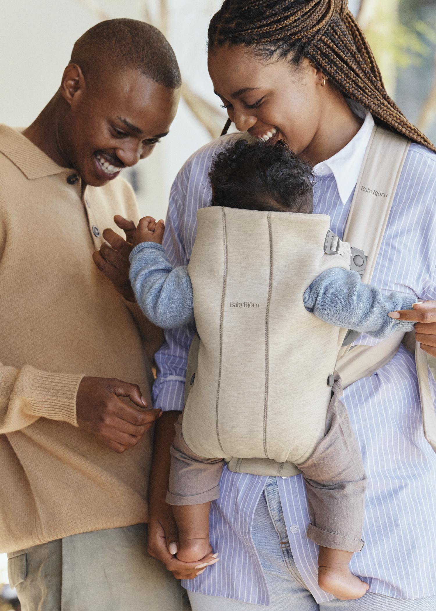 A smiling couple happily looks at their baby in a beige carrier. The father holds the baby's hand while the mother supports the carrier.