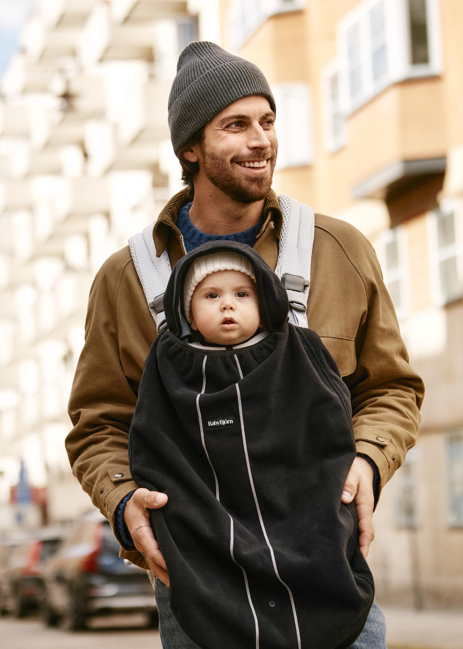 A man in a beanie carries a baby in a front carrier on a city street. Both are warmly dressed, with buildings and parked cars in the background.