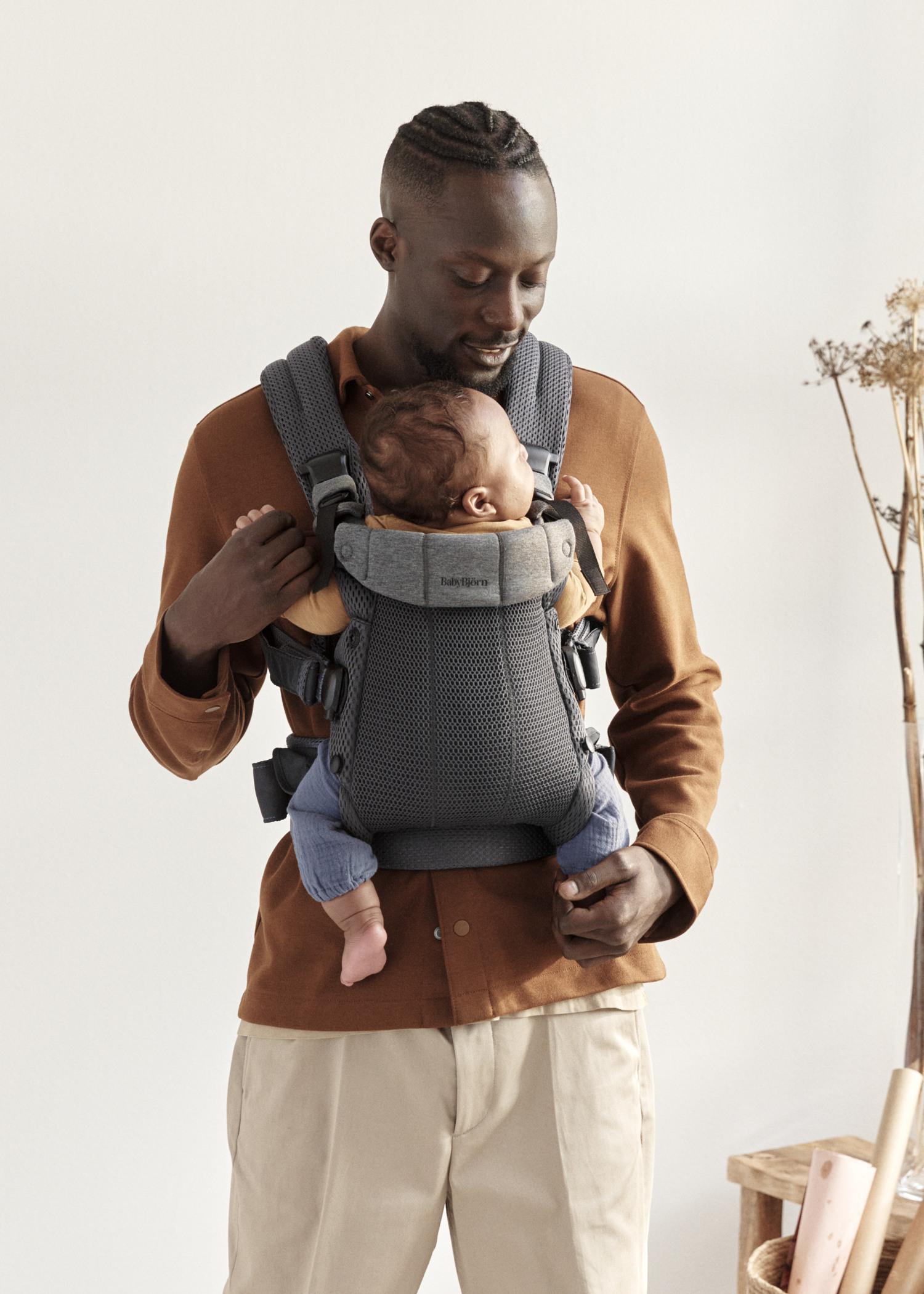 A man with short hair and a beard wears an orange shirt and holds a baby in a gray baby carrier. The baby is sleeping against his chest.