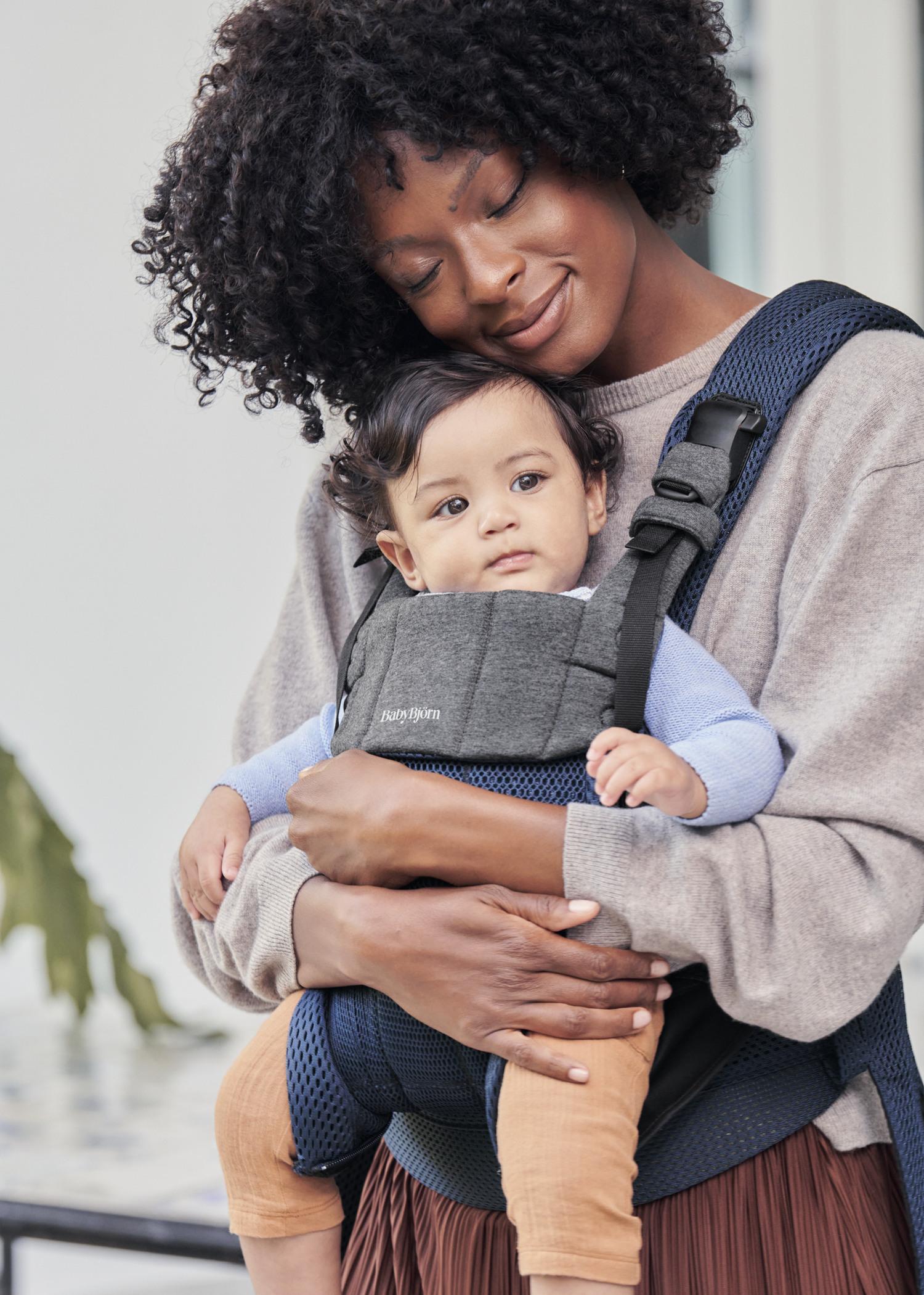 A smiling woman holds a baby in a gray and blue carrier, gently leaning her head on the baby's head. The baby looks curiously at the camera.