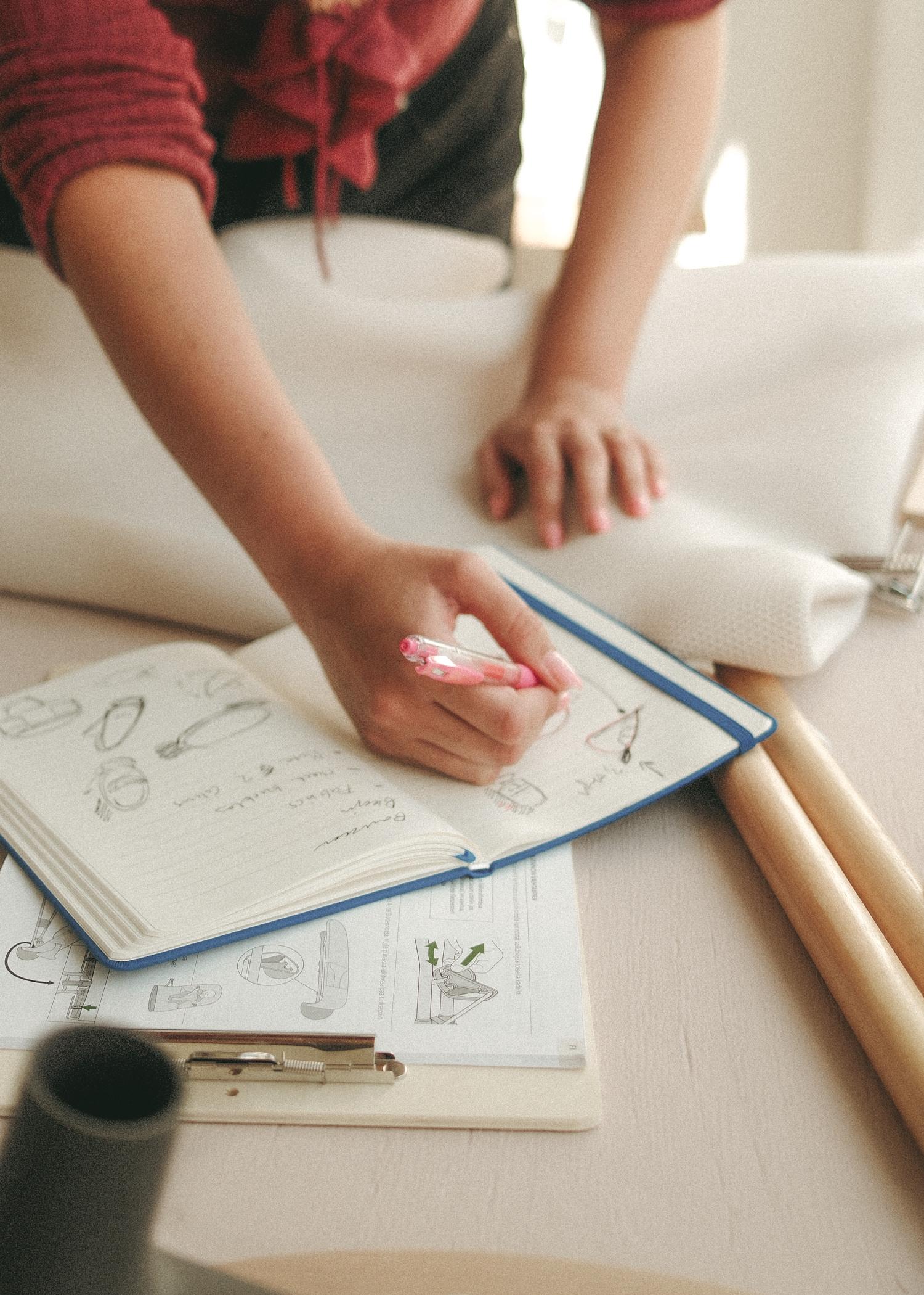 A person drawing sketches and writing notes in a notebook with a pink pen on a table with fabric and a clipboard.
