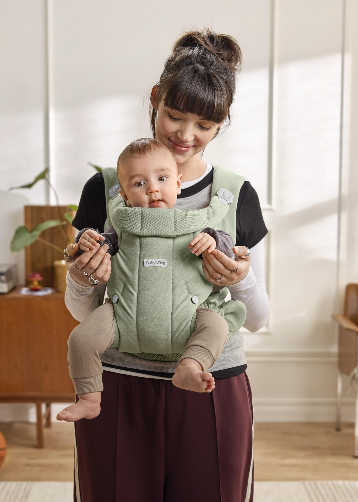 A woman smiles while carrying a baby in a green baby carrier indoors, with a plant and wooden furniture in the background.