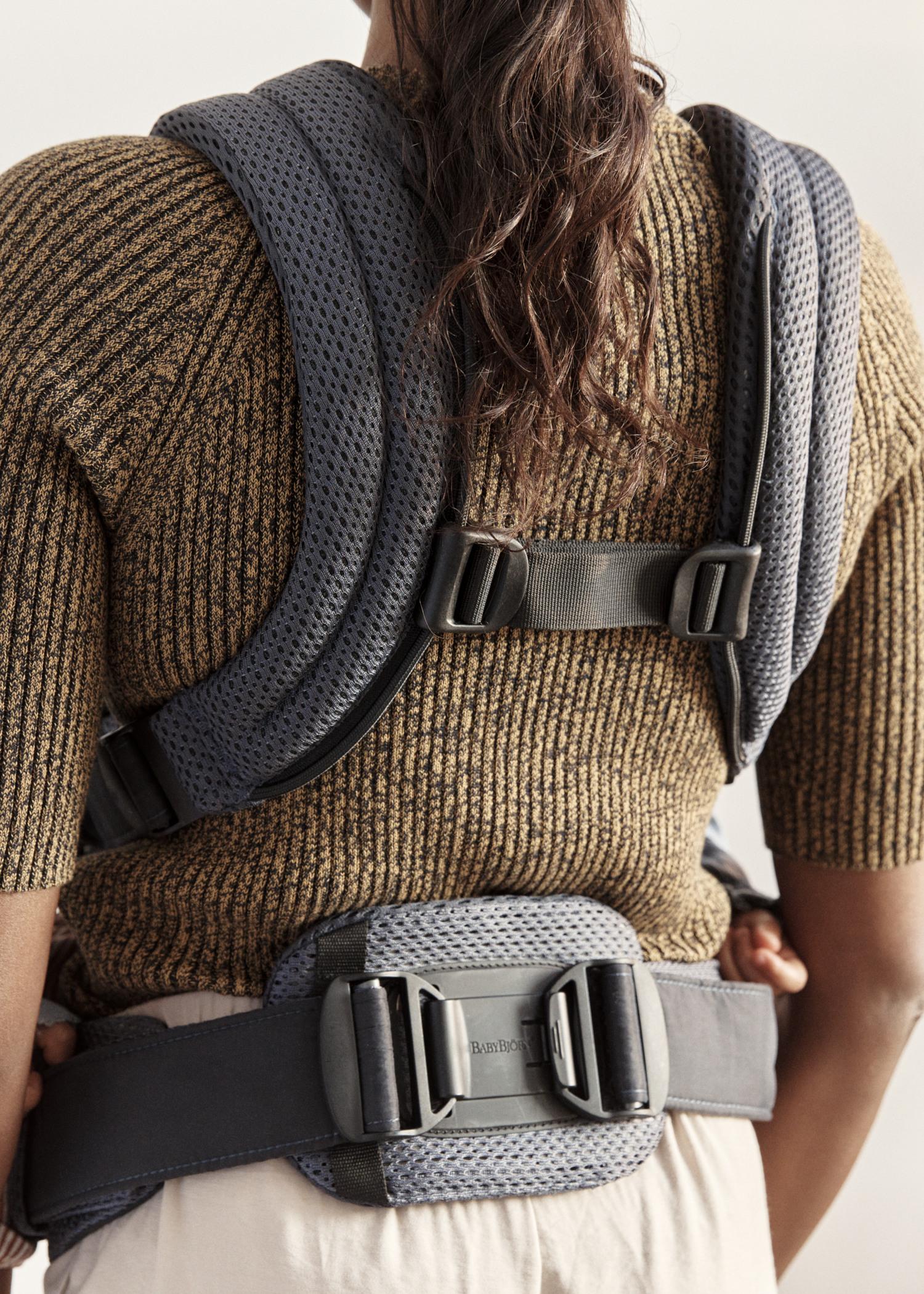 Person wearing a brown sweater and a gray ergonomic baby carrier, viewed from the back, with adjustable straps and buckles.