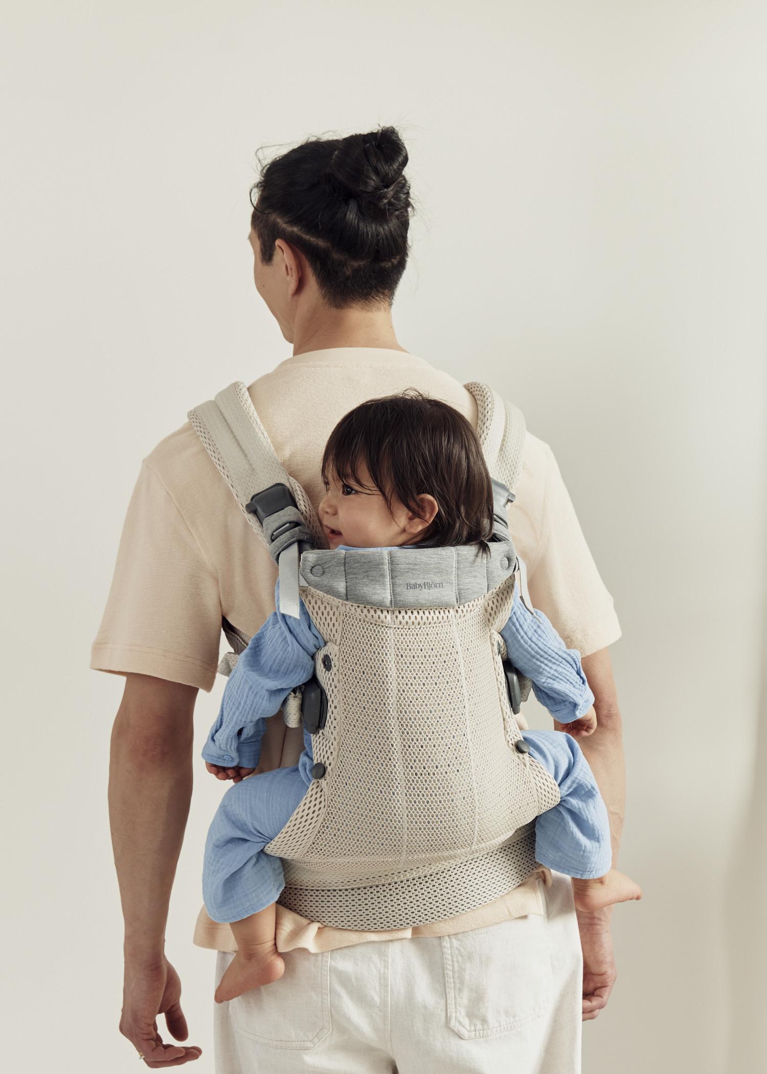 An adult wearing a beige baby carrier holds a child dressed in blue, secured to their back. The adult is facing away, standing against a light background.
