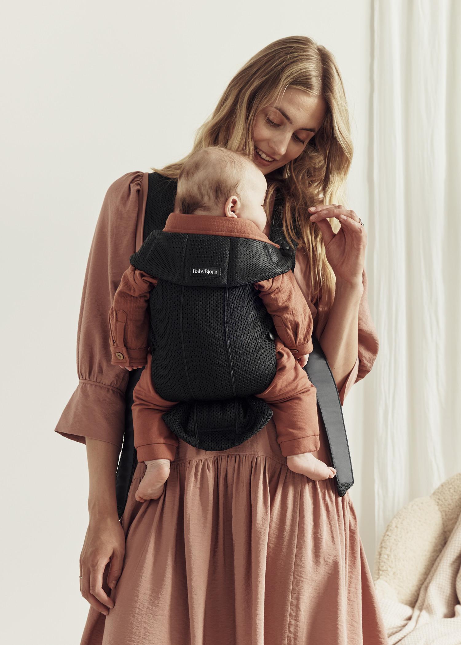 A woman in a pink dress holds a baby in a black carrier. The baby is wearing a rust-colored outfit and is sleeping contently against her chest.