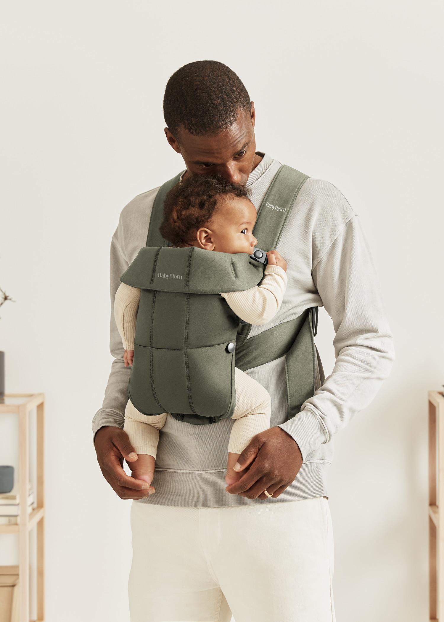A man stands holding a baby in a green baby carrier, both dressed casually; the man gazes affectionately at the baby.