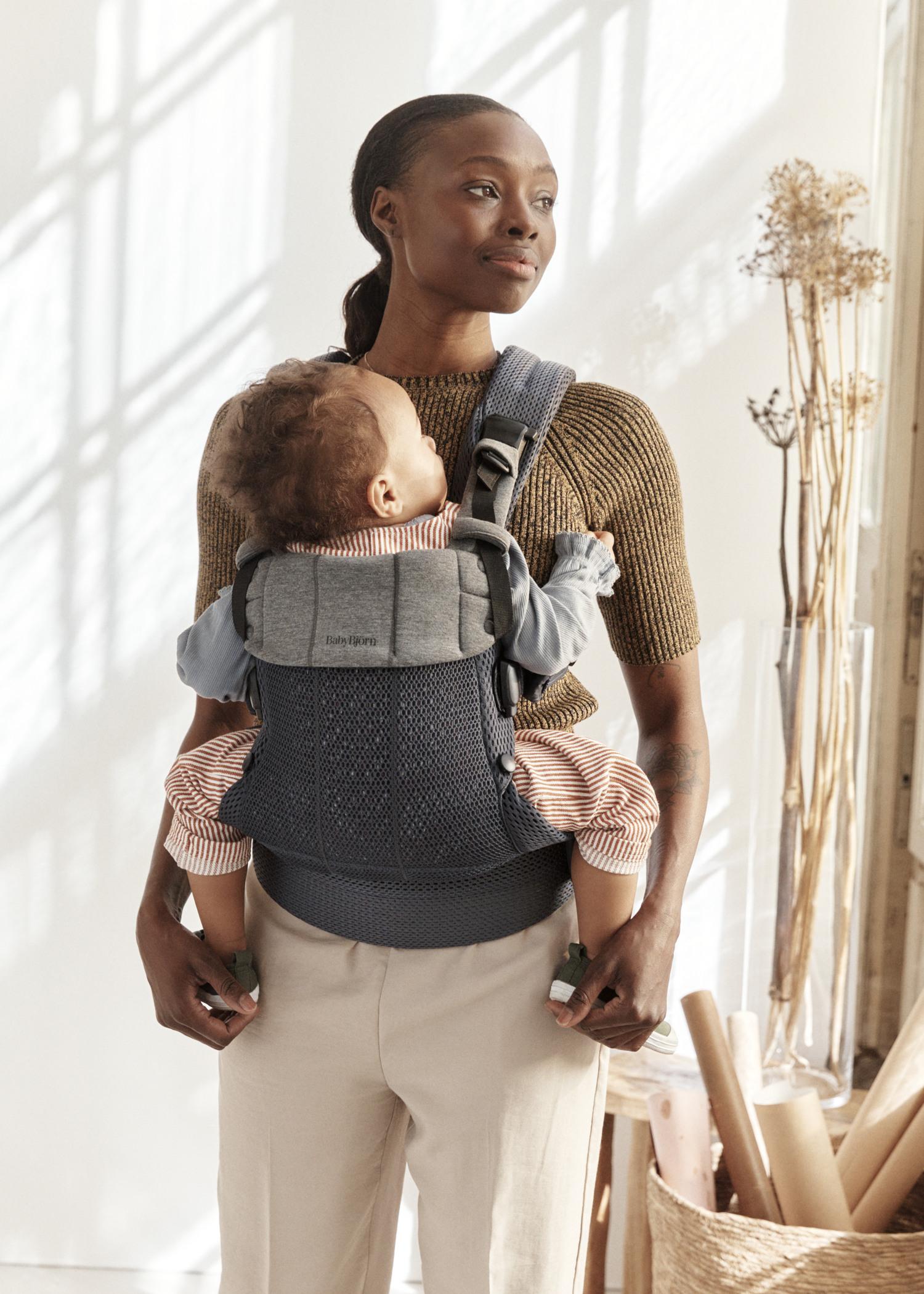 A woman stands near a sunlit window wearing a brown sweater, holding a baby in a carrier on her chest, with dried flowers and rolled papers nearby.