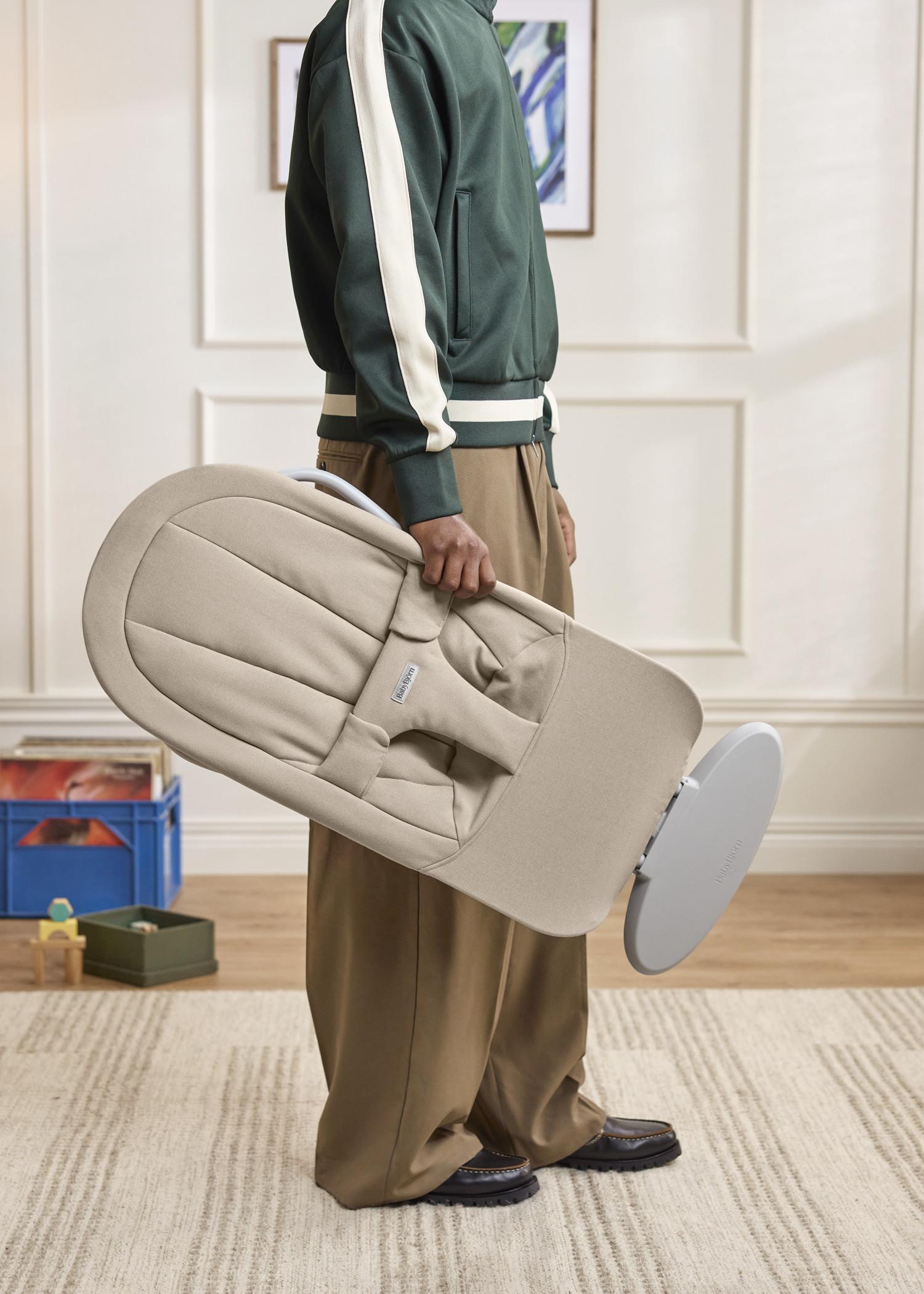 Person in a green and white jacket carrying a beige baby bouncer in a cozy room with toys on shelves.