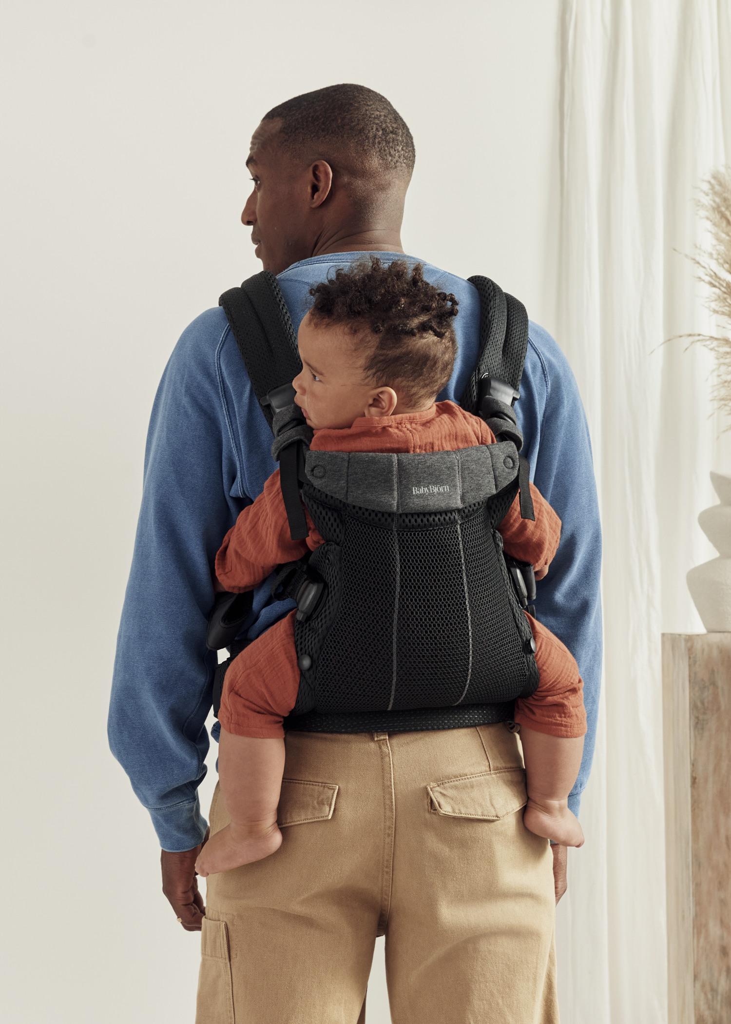 A person wearing a blue sweater carries a baby in a black baby carrier on their back, standing in a light-filled room with a plant.