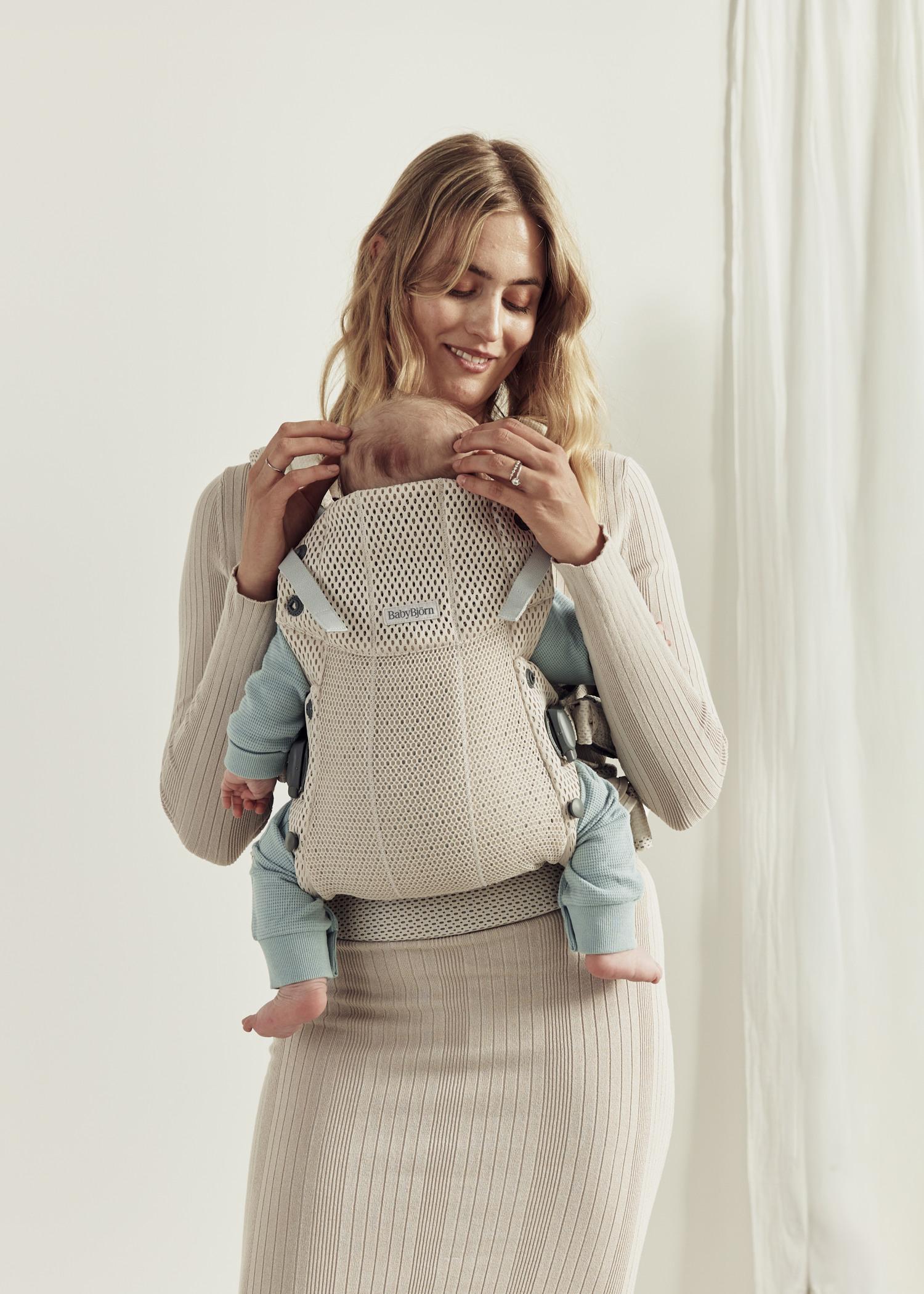 A woman smiles while holding a baby in a beige carrier. She wears a ribbed dress, and the baby is in a light blue outfit. A curtain is in the background.