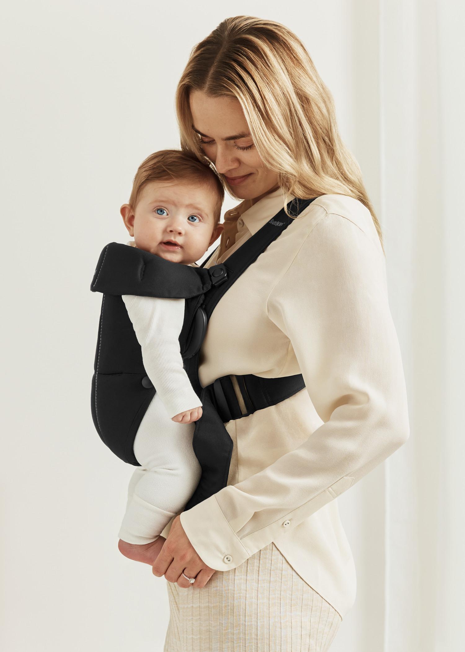 A woman wearing a beige blouse holds an infant in a black baby carrier. The baby, dressed in white, looks forward, and both appear content.