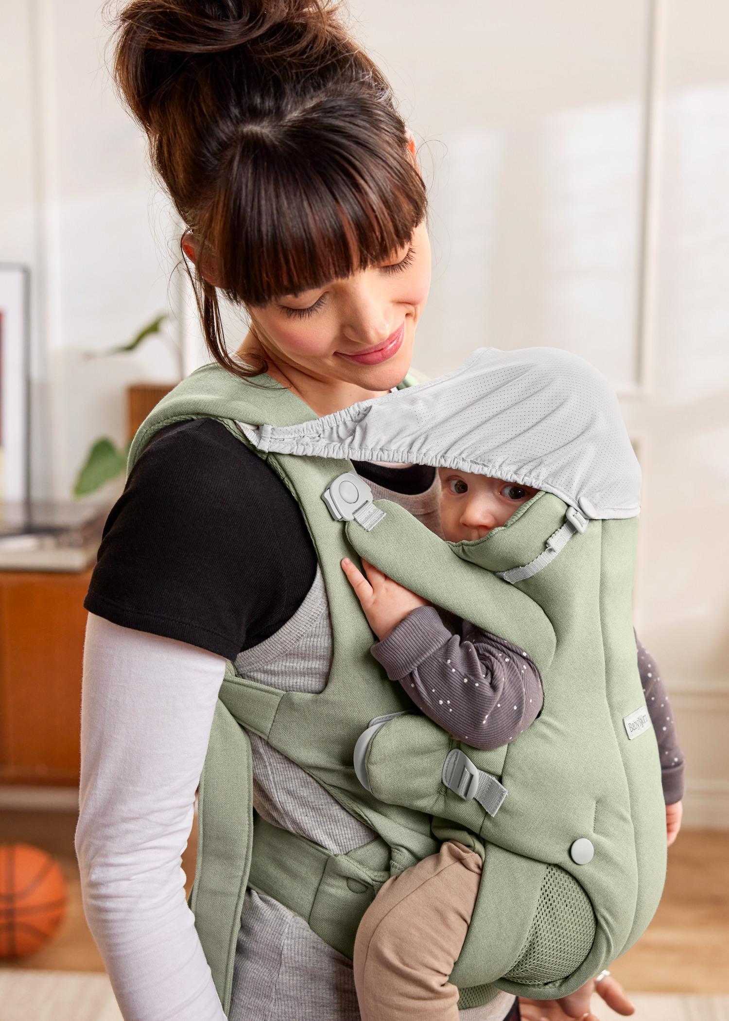 A woman smiles at a baby in a green carrier with a hood, as they stand in a cozy, sunlit room.