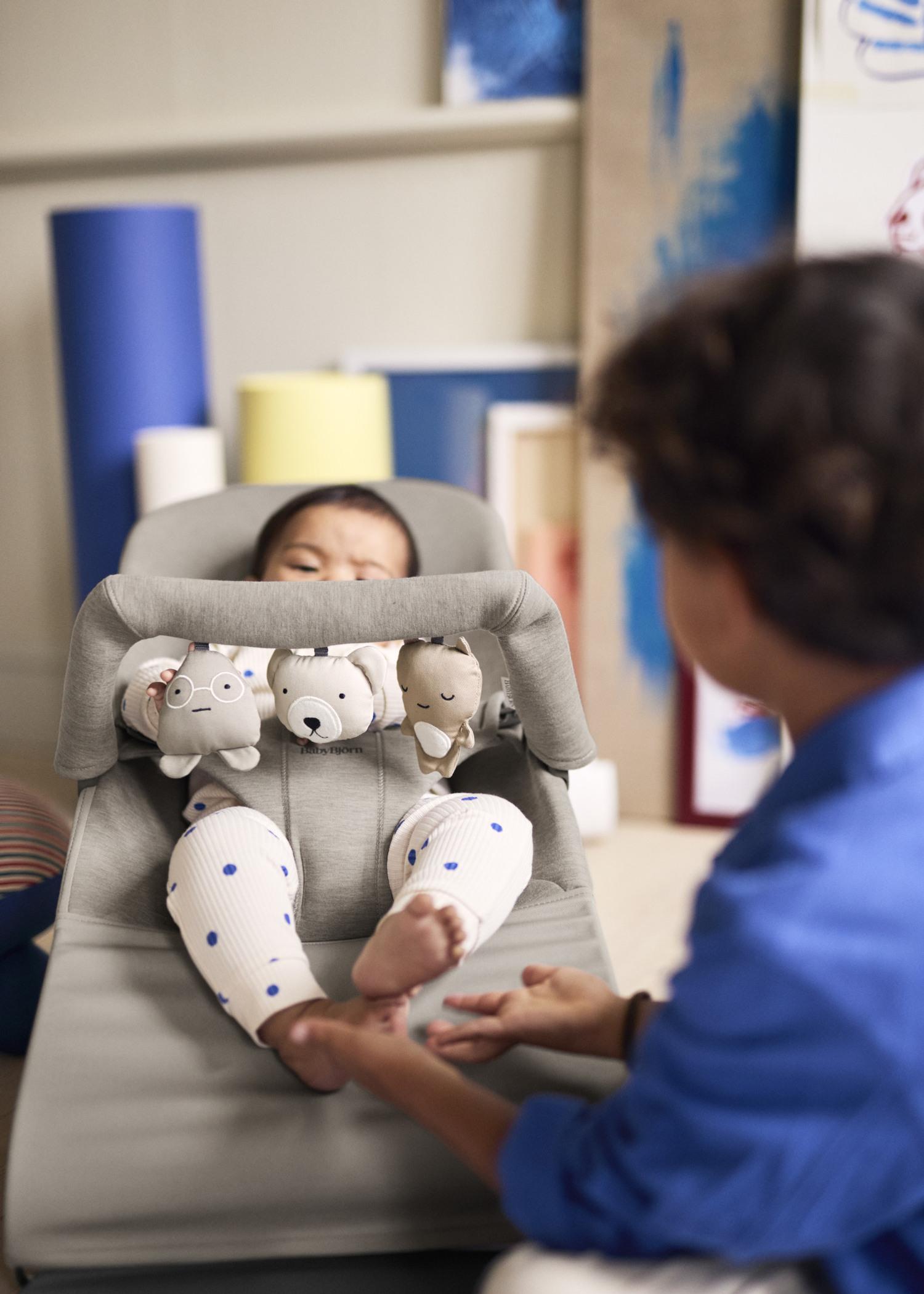 Baby lying in a gray bouncer with hanging animal toys, wearing white pants with blue polka dots. An adult in a blue shirt holds the baby's feet.