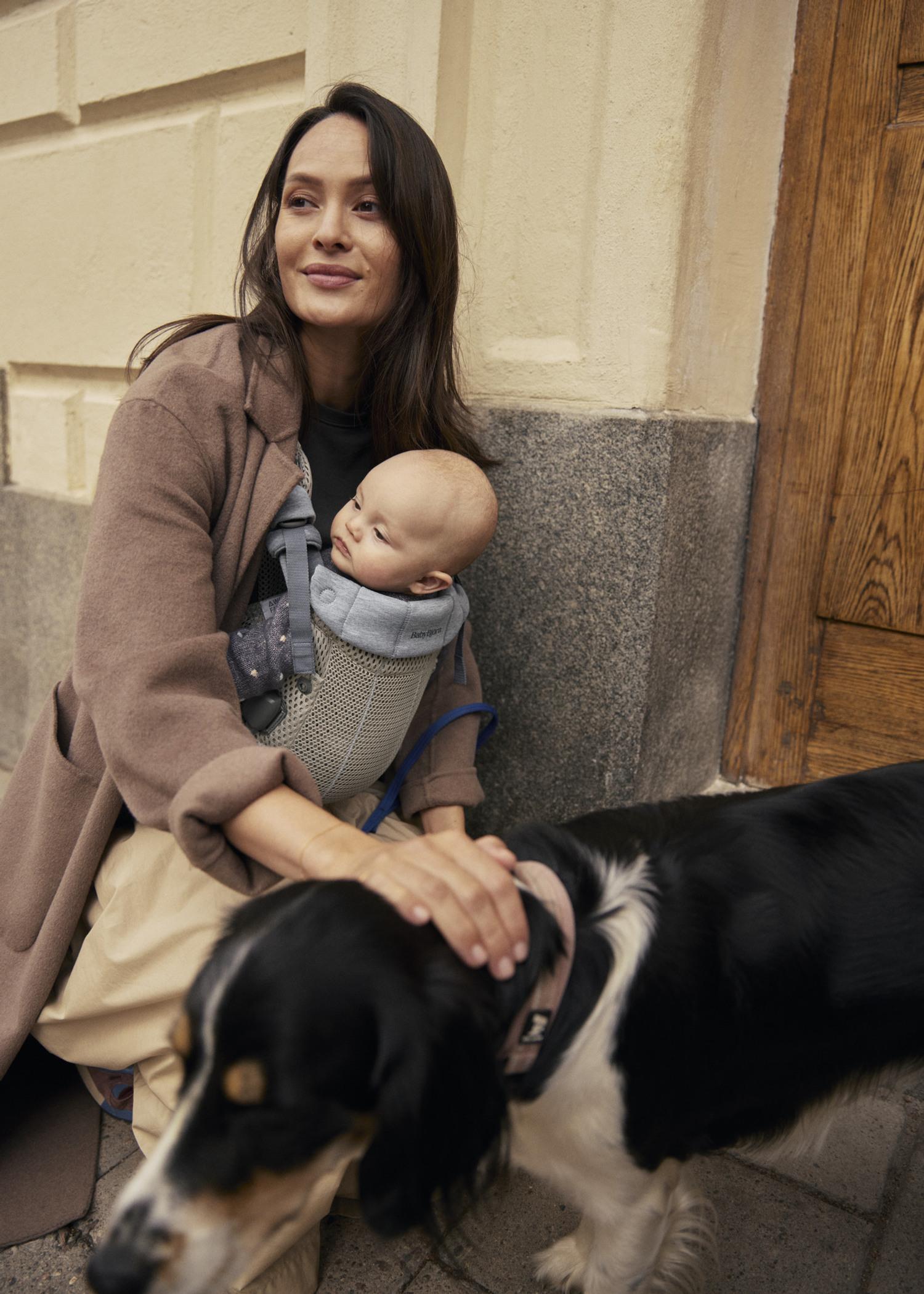 Woman sitting with a baby in a beige baby carrier and a dog beside her, in front of a building with a wooden door.