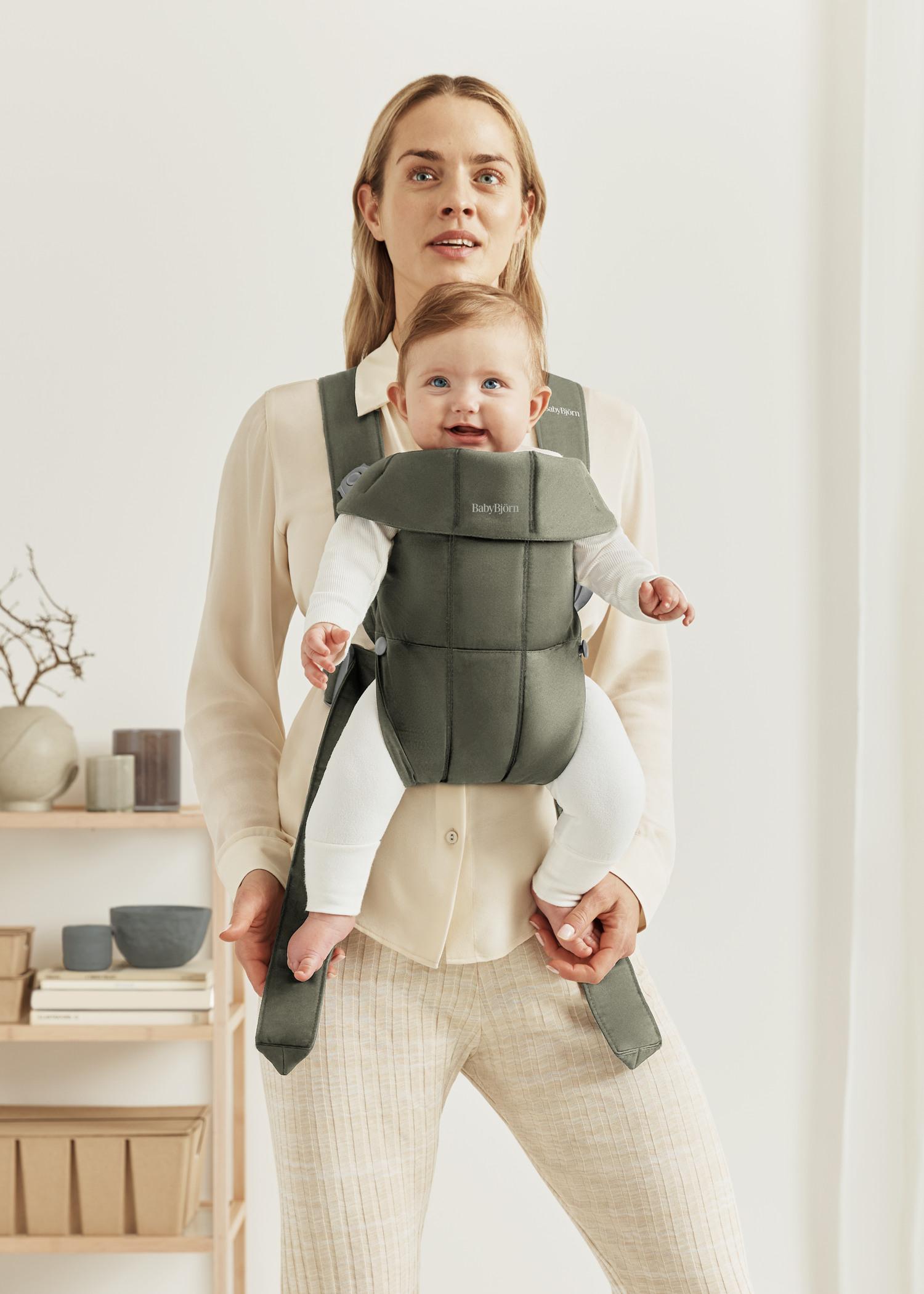 A woman wearing a light blouse and pants carrying a smiling baby in a green baby carrier. The background shows shelves with decor items.
