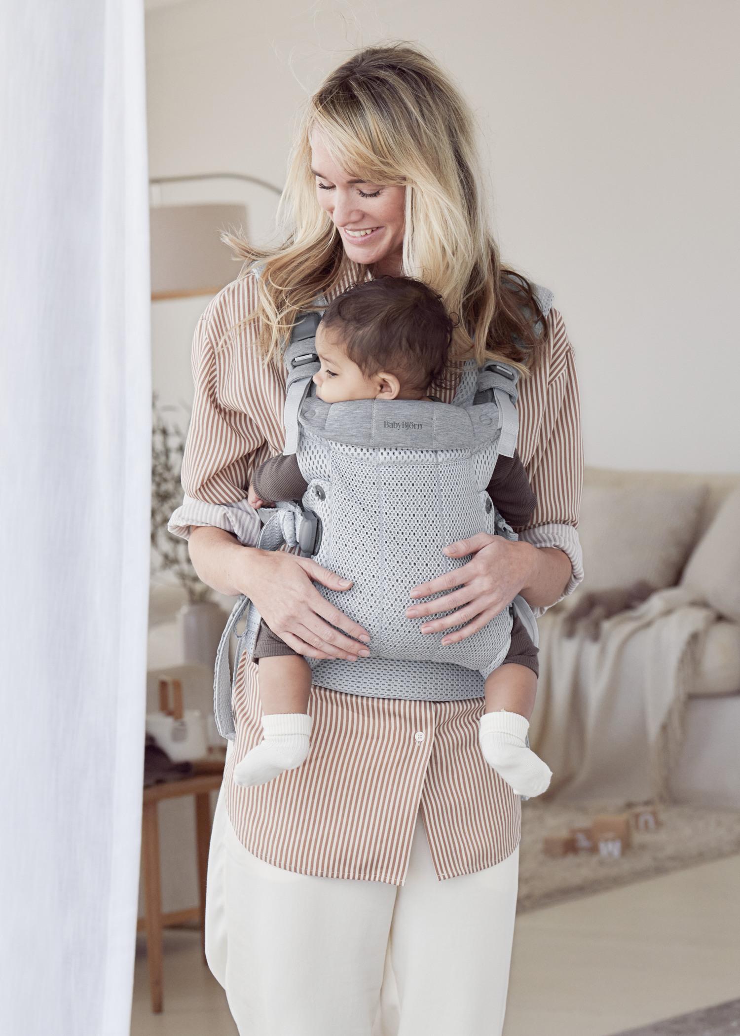 A woman smiles while holding a baby in a gray carrier in a cozy, softly-lit room with neutral colors.