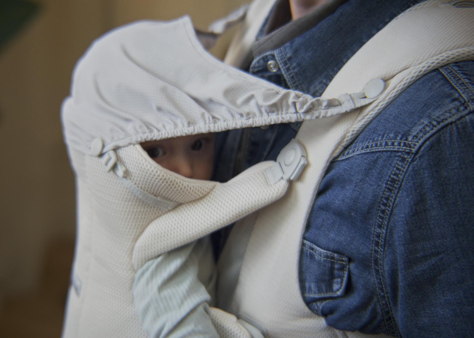 A baby peeks out from a cream-colored baby carrier, snuggled against a person wearing a denim shirt.