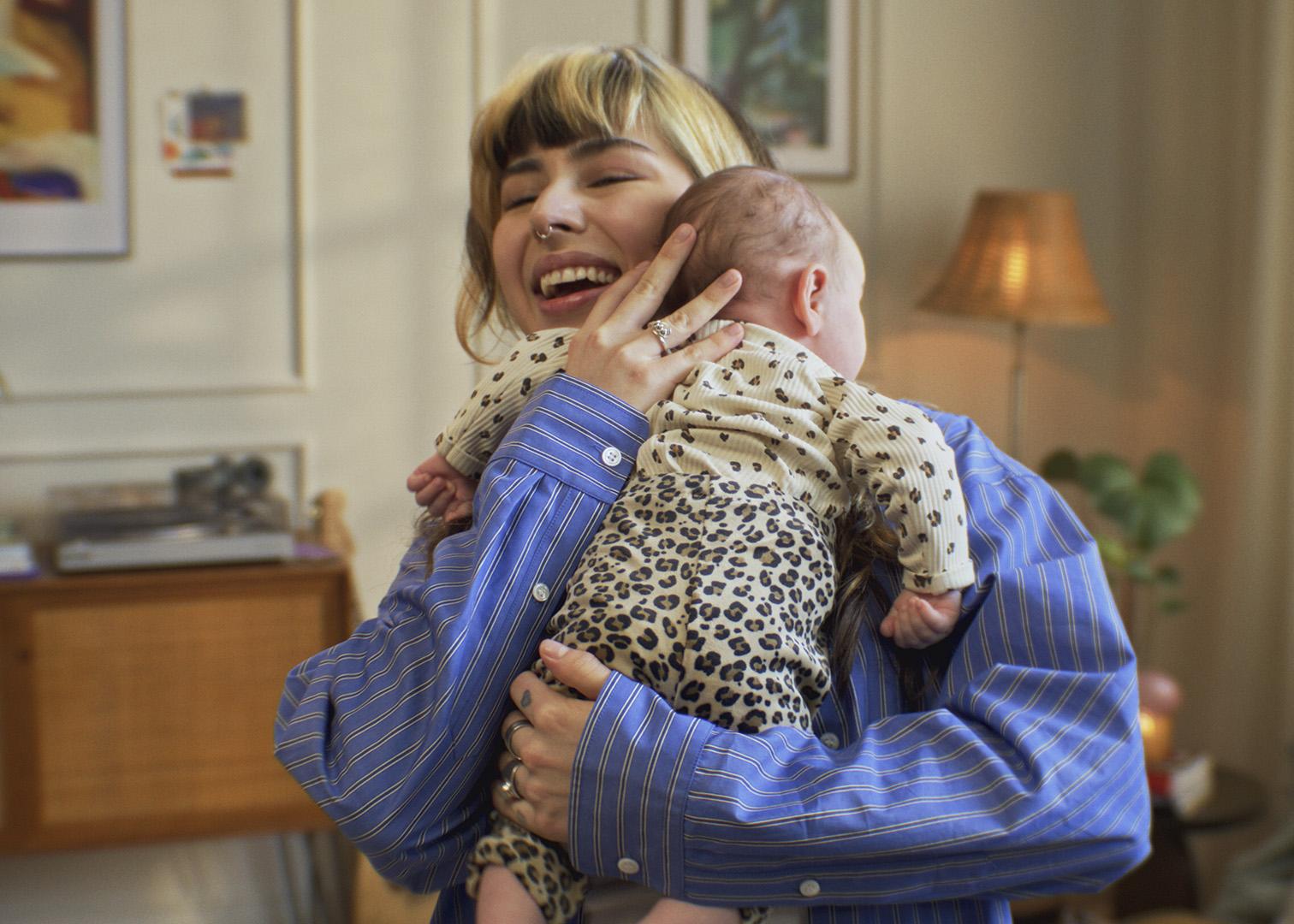 Woman in a blue shirt smiling while holding a baby in leopard print pajamas, standing in a warmly lit room with lamps and wall art.