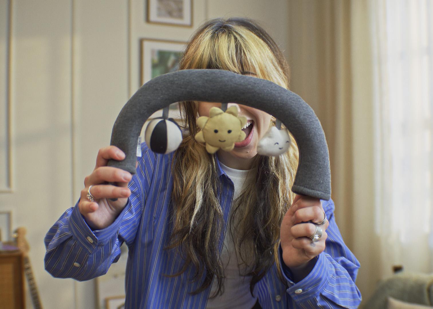 Person holding a plush gray toy arch with hanging stuffed animals in a cozy room.