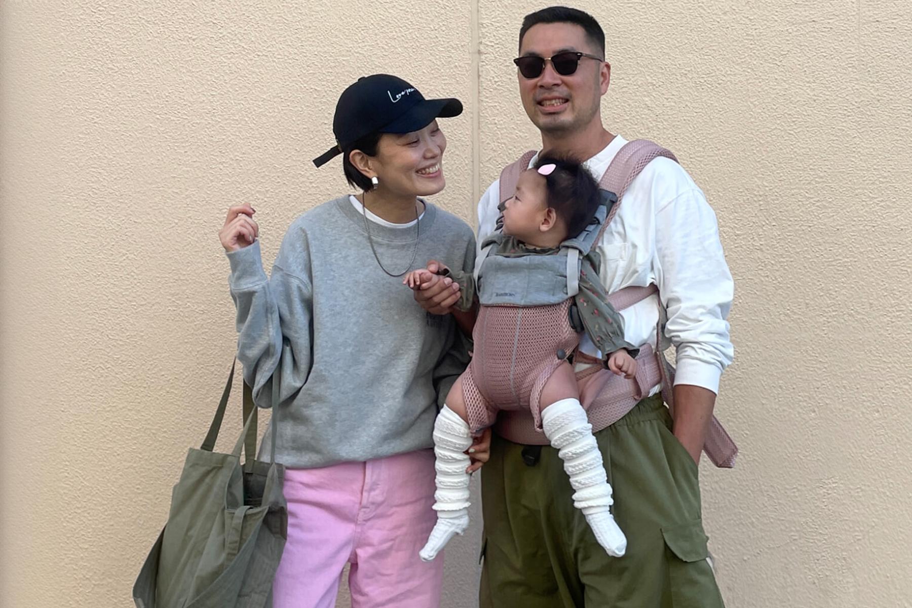 A smiling couple stands together; the man holds a baby in a carrier. The woman wears a cap and casual clothes. The baby wears a headband.