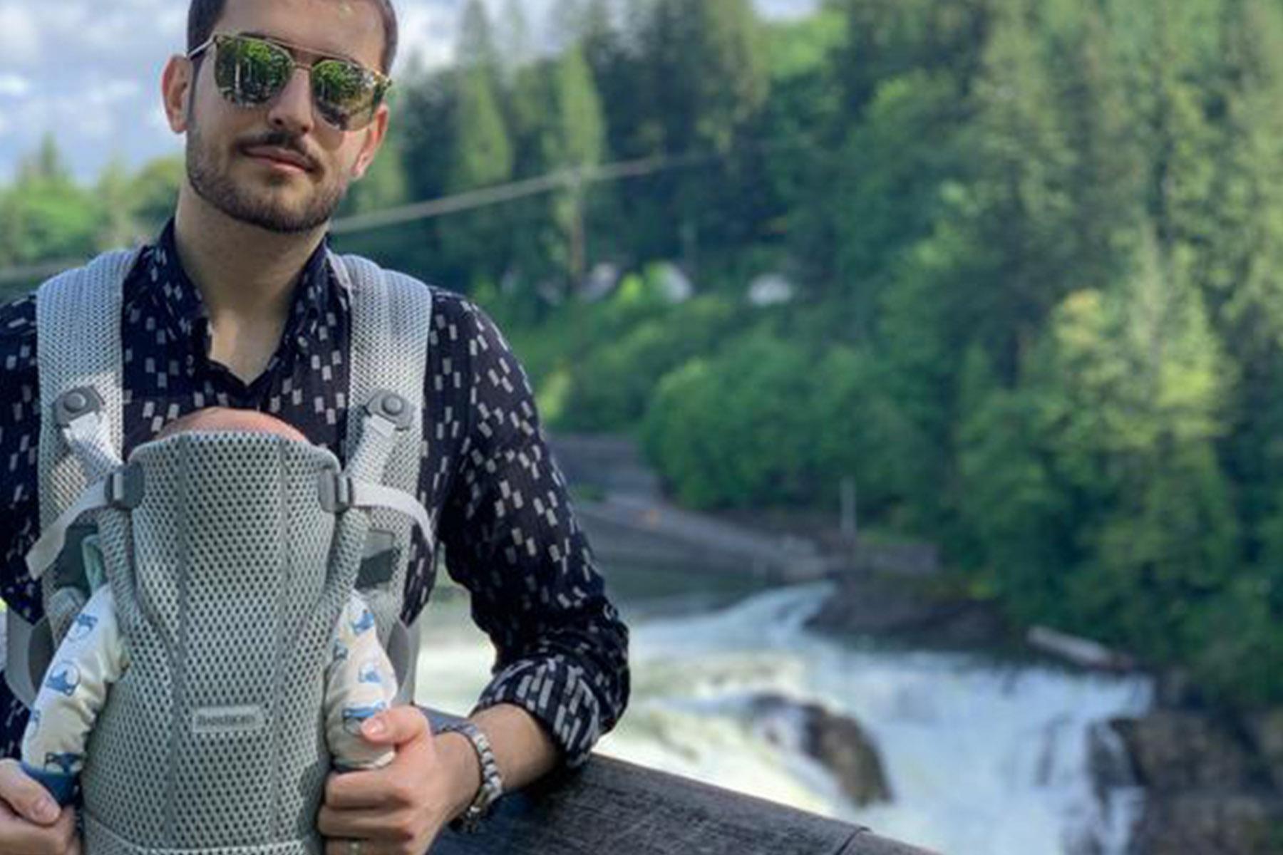Man in sunglasses holding an infant in a front baby carrier on a wooden railing overlooking a waterfall and lush forest.