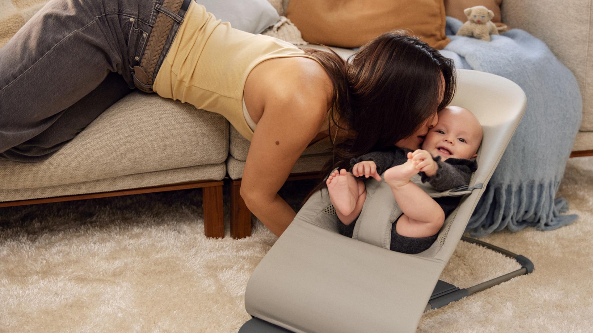 A woman playfully kisses a smiling baby seated in a gray baby bouncer on a beige carpet near a sofa.