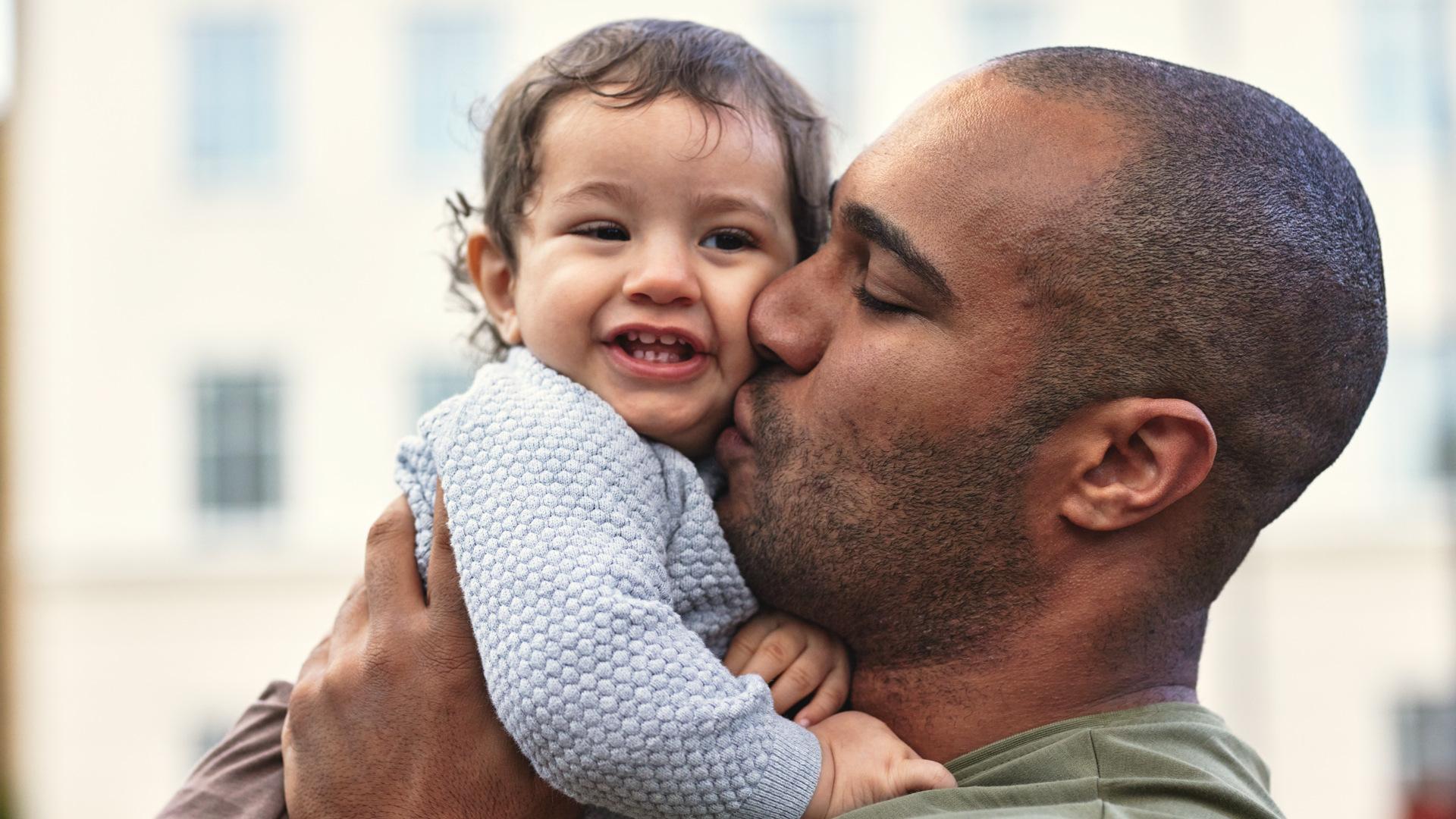 A man lovingly kisses a smiling baby on the cheek while holding the child in his arms outside.