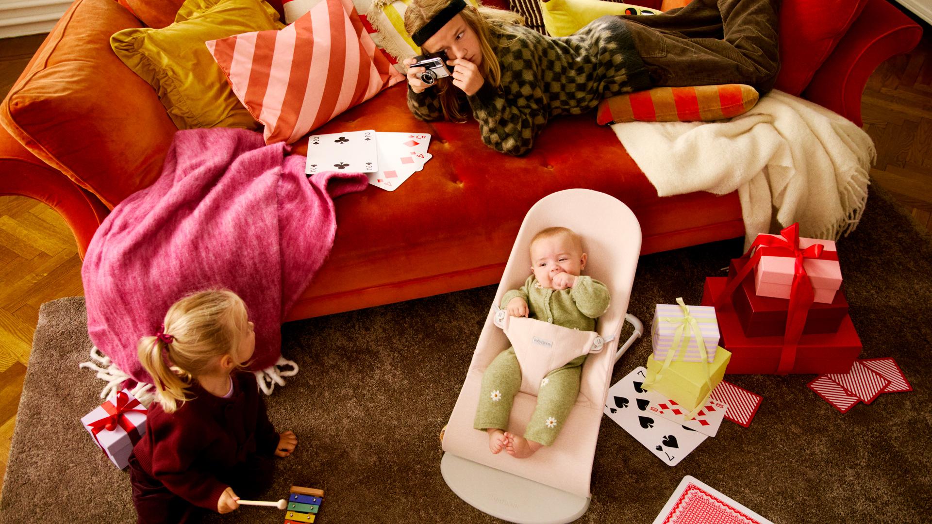 A cozy living room scene with a baby in a BabyBjörn bouncer, a child playing, and an adult taking photos. Gifts and playing cards are scattered around.