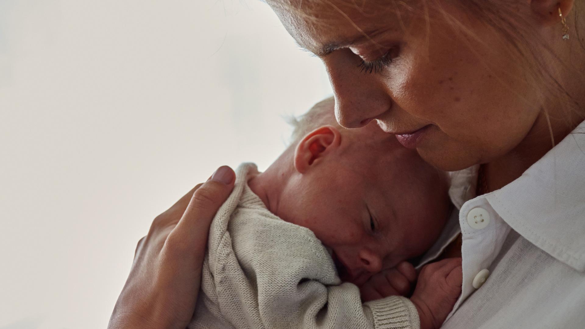 A woman gently cradles a sleeping baby against her chest, her face tenderly touching the baby's head, in a peaceful and intimate moment.