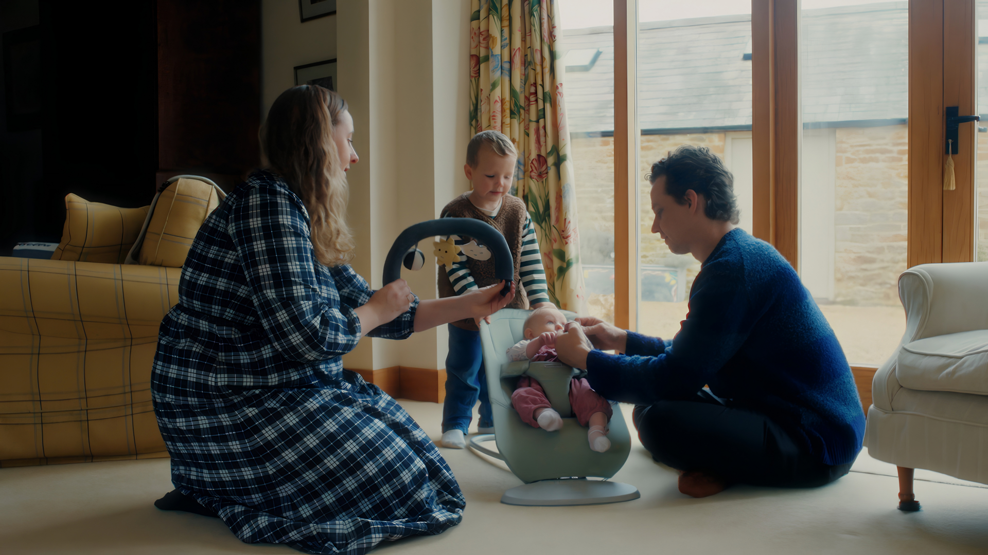 A family gathered around a baby in a bouncer, with a woman holding a toy and a man adjusting the baby, while a child stands nearby.