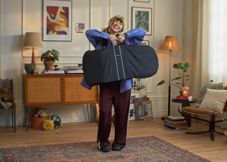 A person stands in a cozy living room, smiling and holding a large black transport bag for bouncer. The room features stylish decor and plants.