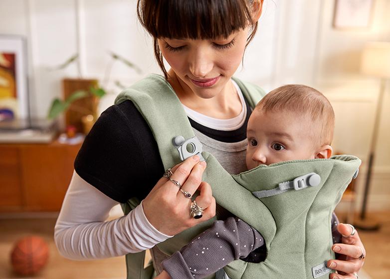 A woman wearing a baby carrier Hold looks down at a baby snugly seated inside it, both indoors with a warm ambiance.