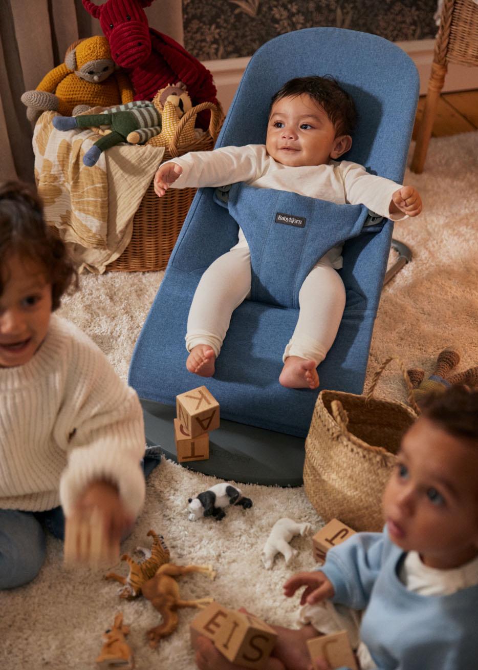 Baby in a blue bouncer chair, surrounded by two toddlers playing with wooden blocks and toy animals on a soft carpet.